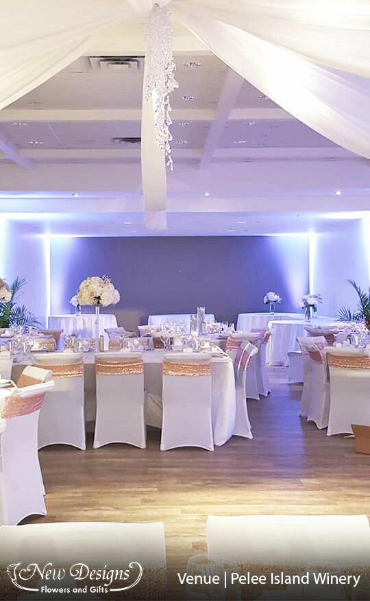 A hall is decorated with a gorgeous draped white ceiling, tall centerpieces of Hydrangeas and roses compact atop a tall clear fluted vase run through with twinkle lights. And ambient lighting spikes upwards in the corners of the room.